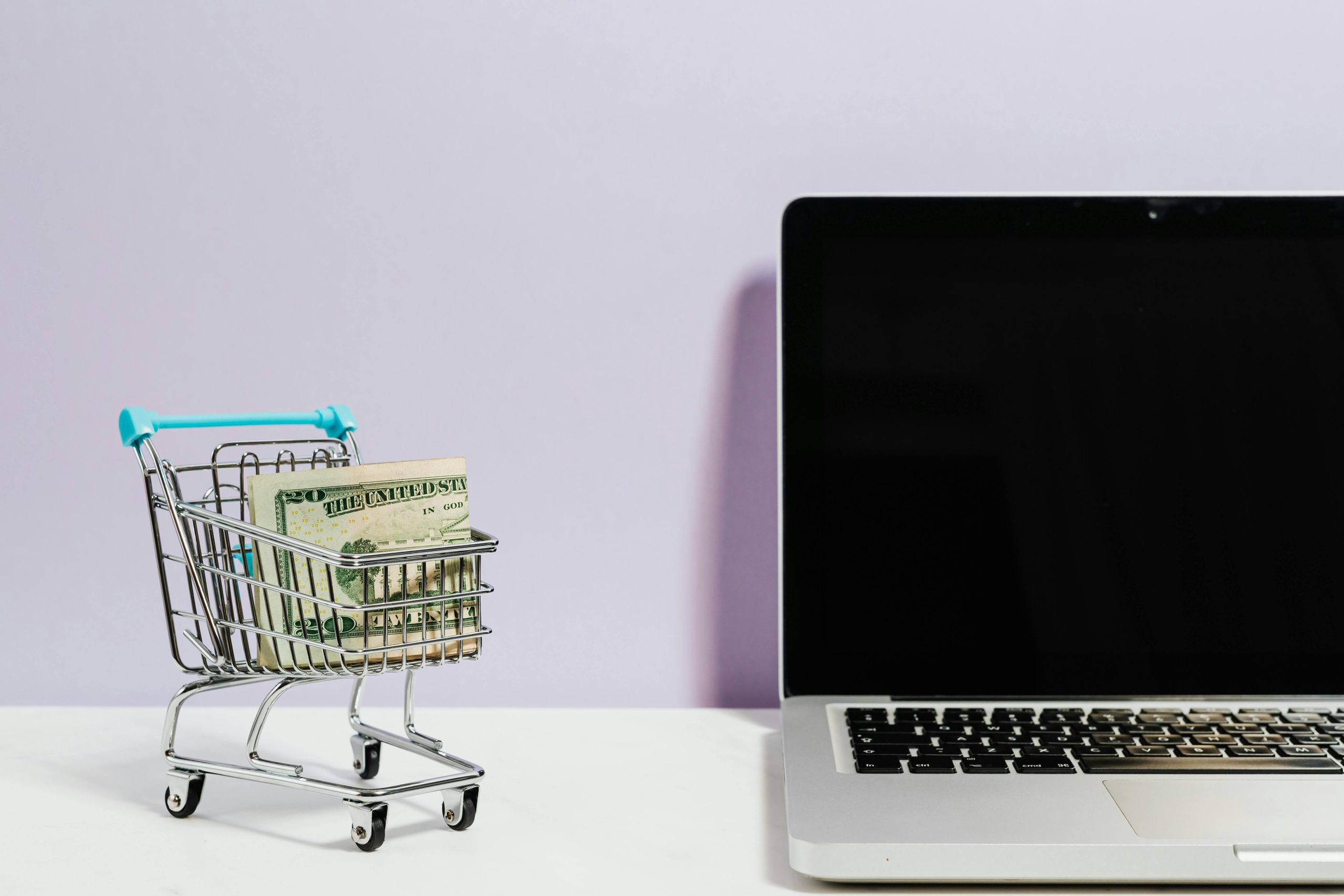 bundle of cash in a miniature shopping cart, next to a laptop